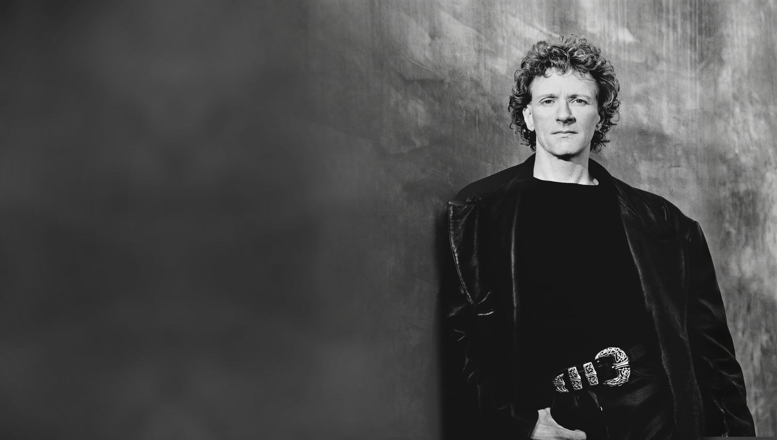 Rama in a black leather coat against a mottled grey studio wall, black and white portrait by Greg Gorman