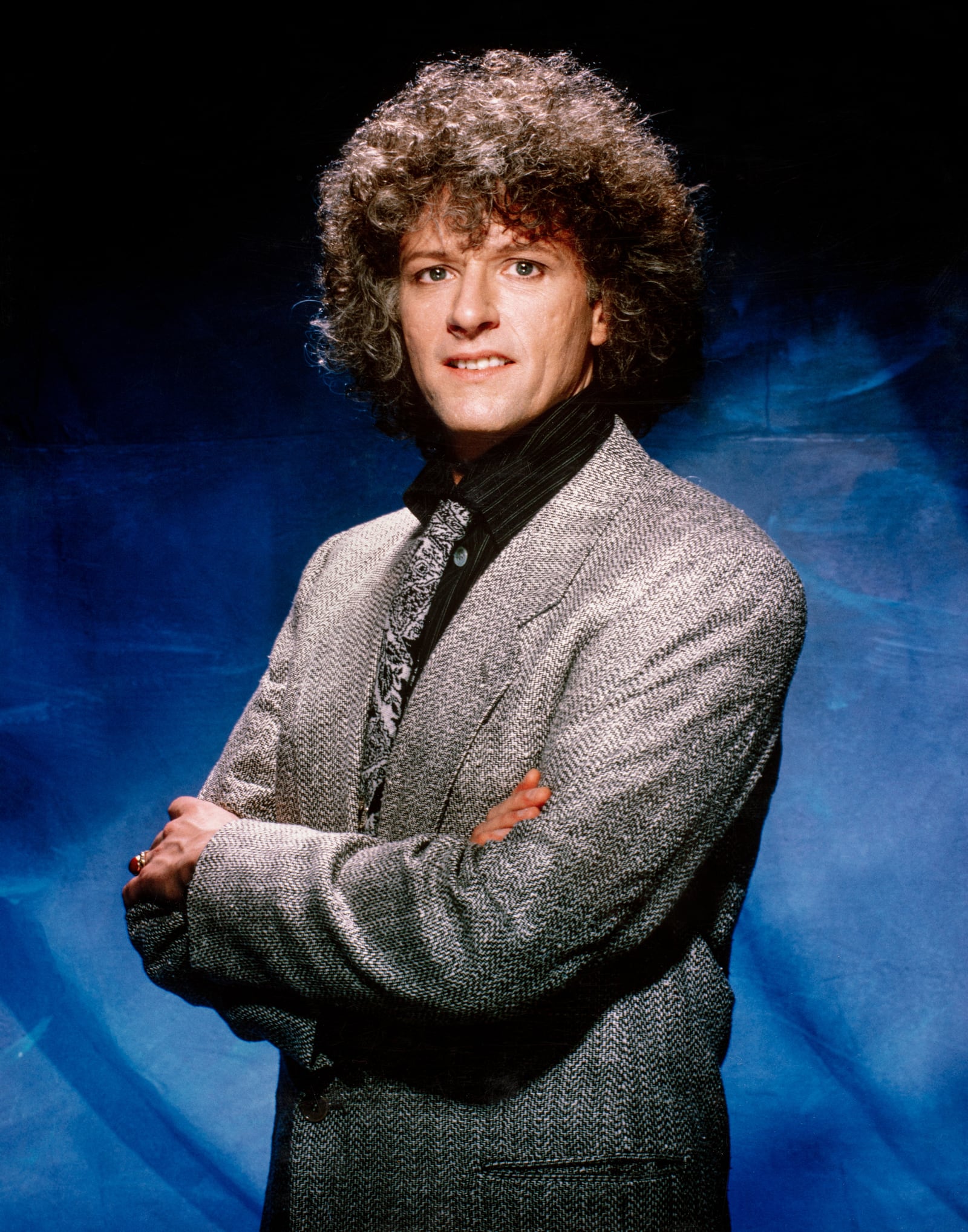 Rama in a grey tweed jacket with arms crossed against a blue-lit backdrop, 1980s studio portrait